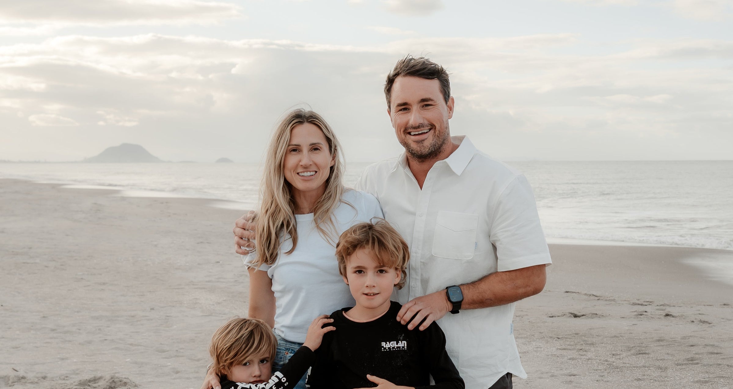 Dr. Clive Price, founder of True Proactive, a NZ based bowl cancer screening company, and his family posing together on a sandy beach with the ocean and cloudy sky in the background. The father wears a white short-sleeve button-up shirt, the mother wears a white top, and the two boys wear black shirts.